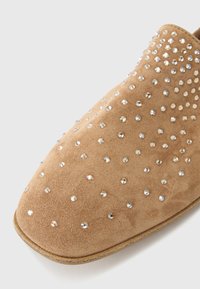 Καστανό μπεζ παπούτσι τύπου slip-on από σουέντ, διακοσμημένο με διάφανα στρας, με στρογγυλερή μύτη και διακριτή υφή. Μαλακή επιφάνεια με λείο φινίρισμα.