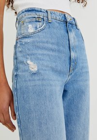 Jeans de tiro alto de color azul claro, hechos de denim, con parches deshilachados, diseño tradicional de cinco bolsillos y suturas amarillas sutiles.