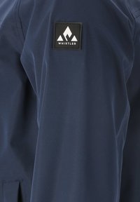 Marineblå, vattentät jacka med en slät textur; har en framträdande svart fyrkantig patch som visar "WHISTLER"-logotypen och bergdesign.