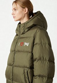 Olijfgroene gewatteerde jas met capuchon, voorzien van een geborduurd "I ♥ YOU"-ontwerp in rood en wit op de borst, en een ritssluiting aan de voorkant.