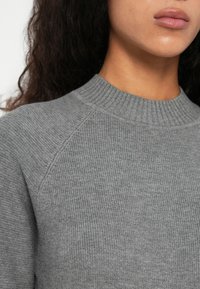 Mlad človek z temnimi kodrastimi lasmi, ki nosi sivo rebrasto pleteno pulover z visokim okroglim izrezom in raglan rokavi.