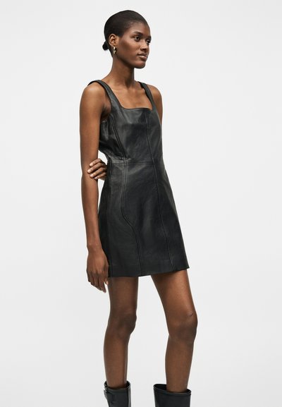 Γυναίκα με σκουρόχρωμο δέρμα και κοντά μαλλιά που φοράει ένα μαύρο, δερμάτινο, μίνι φόρεμα χωρίς μανίκια και μαύρες μπότες, στέκεται με το ένα χέρι σταυρωμένο πίσω της.