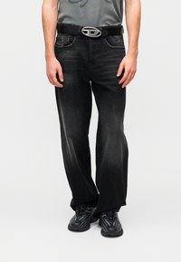 Μαύρο τζιν από denim με άνετη εφαρμογή, διαθέτει μπροστινές τσέπες, ορατό λογότυπο στη αγκράφα της ζώνης και διακριτικές ραβδώσεις στο ύφασμα.