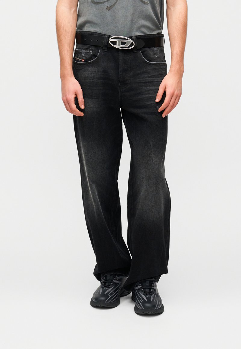 Μαύρο τζιν από denim με άνετη εφαρμογή, διαθέτει μπροστινές τσέπες, ορατό λογότυπο στη αγκράφα της ζώνης και διακριτικές ραβδώσεις στο ύφασμα.