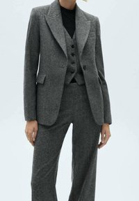 Grijze wollen pak met een op maat gemaakte blazer met pieklapels en een vest met drie knopen, gecombineerd met een broek met wijdere pijpen, alles in een zachte textuur.