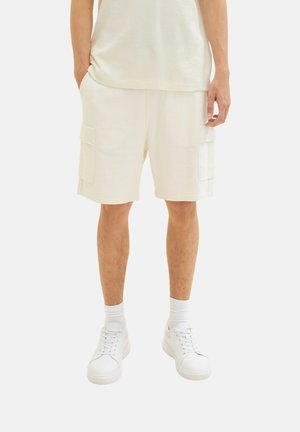 Cremefarbene Cargo-Shorts mit zwei Seitentaschen, gefertigt aus einem strukturierten Stoff. Kombiniert mit weißen Sneakers und Socken.
