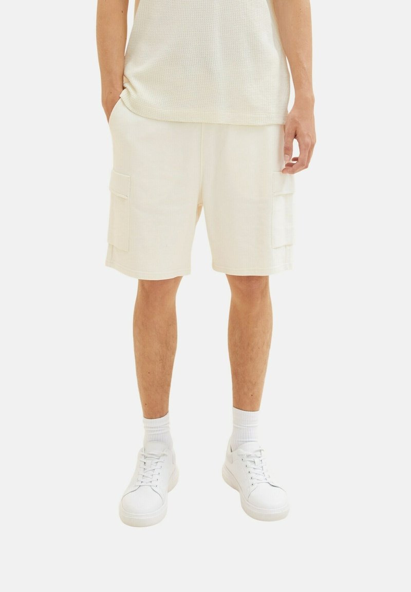 Shorts cargo crème avec deux poches latérales, confectionnés dans un tissu texturé. Associés à des baskets blanches et des chaussettes.