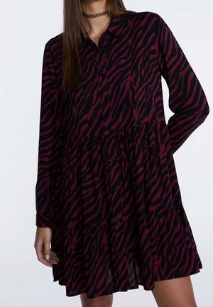 Vrouw die een jurk met lange mouwen en knopen draagt, met een zwart en donkerrood zebraprint, staand tegen een effen lichte achtergrond.