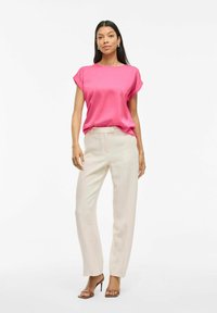 Blusa rosa di satin a maniche corte abbinata a pantaloni in lino beige chiaro. Il modello indossa sandali con tacco alto color cammello dal design minimalista.
