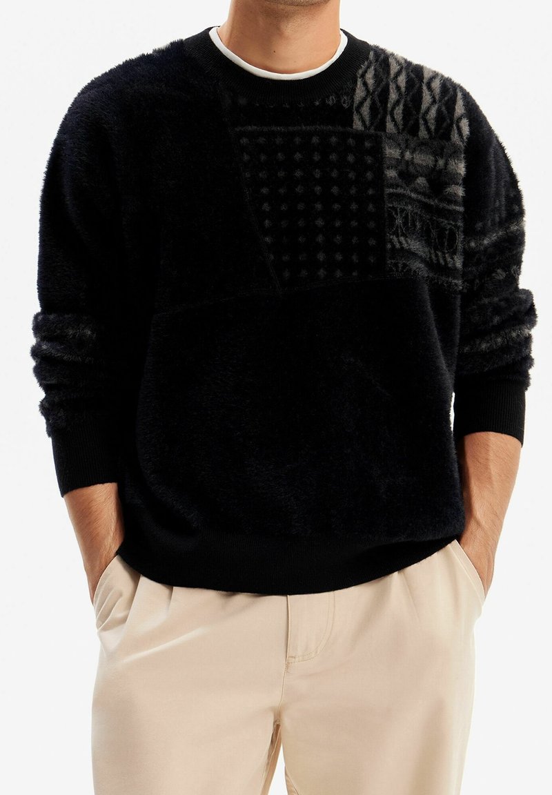 Pull noir en tissu duveteux avec motifs géométriques en patch texturé, porté par une personne ayant les mains dans les poches d'un pantalon beige, avec un col rond blanc visible.