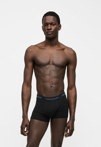 Uomo con pelle scura e corporatura atletica in piedi su uno sfondo semplice, che indossa boxer neri Tommy Hilfiger.