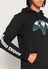 Sort hættejakke med en kængurulomme, prydet med et turkist geometrisk logo og hvid tekst, der læser "ADIDAS ORIGINALS" ned ad ærmet.