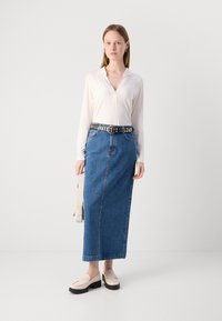 Balta ilgomis rankovėmis marškiniai, aukštu liemeniu mėlyna džinsinė sijonas, juodas dekoruotas diržas ir smėlio spalvos platformos loafersai. Neutralus fonas.