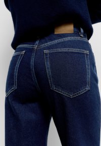 Takakuva henkilöstä, joka käyttää tummansinisiä farkkuja ja tummansinistä neulepaitaa, näkyvissä takataskut ja ruskea nahkainen vyötärönauhassa oleva merkki.