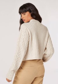 Creme kabelstrik sweater med en cropped design og ribbede manchetter, der har en tekstureret ryg. Sammen med lysebrune tætsiddende bukser.