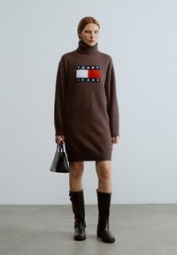 Καφέ πλεκτό φόρεμα με ψηλό γιακά, που διαθέτει πολύχρωμο λογότυπο. Το μοντέλο φοράει μαύρες μπότες που φτάνουν μέχρι το γόνατο και κρατάει μια τσάντα.