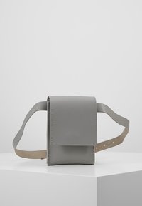 Šedá kožená crossbody taška s klopovým uzávěrem, hladkou texturou, kontrastním hnědým popruhem a viditelnými švy na okrajích.