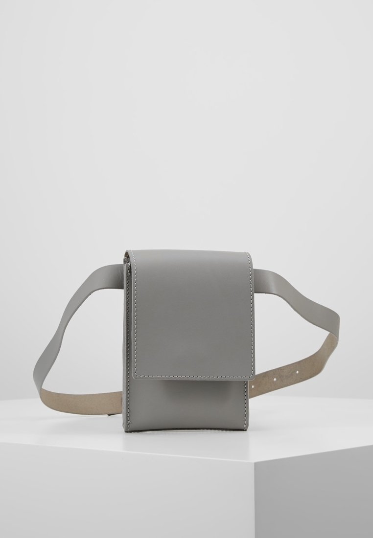 Šedá kožená crossbody taška s klopovým uzávěrem, hladkou texturou, kontrastním hnědým popruhem a viditelnými švy na okrajích.