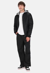 Sort jakke med grå hætte, hvid t-shirt, løse sorte bukser og sorte sneakers med røde detaljer. Glat stof, afslappet pasform.