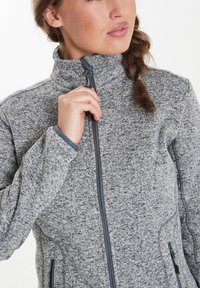 Veste en polaire grise avec un col haut, fermeture éclair sur le devant et poches latérales. Présente un tissu texturé et des coutures contrastées.