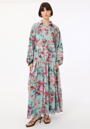 Vrouw draagt een lange, lichtblauwe bloemenjurk met een taillekoord en lange mouwen, gecombineerd met bruine sandalen met hak en bandjes en gouden oorringen.
