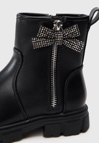 Bota de tobillo de cuero negro con cremallera plateada, decorada con un lazo de pedrería en el lateral y una suela gruesa de goma. Textura lisa.