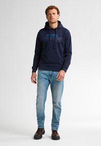 Hanorac bleumarin cu sireturi și grafică frontală, asortat cu blugi de culoare albastru deschis și cizme maro închis. Îmbrăcăminte casual, confortabilă.