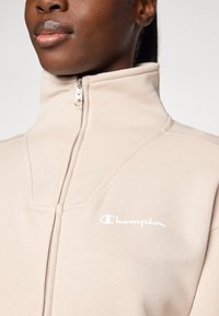 Veste en polaire zippée beige clair avec col haut, présentant un logo "Champion" blanc et des accents de tissu texturé subtils.
