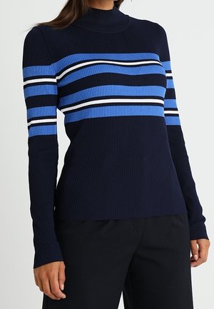 Kvinde iført en marineblå ribstrikket sweater med horisontale blå, hvide og sorte striber over brystet og ærmerne, kombineret med sorte bukser.