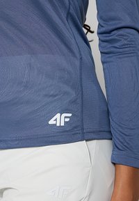 Jasnoniebieska sportowa koszula z długim rękawem, wykonana z teksturowanej tkaniny, z okrągłym dekoltem. Posiada białe logo w lewym dolnym rogu oraz subtelne wzory.