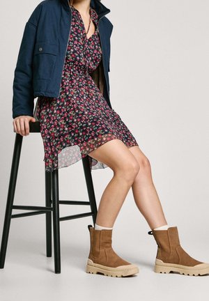 Personne assise sur un tabouret noir, portant une robe fleurie, une veste marine et des bottes en daim beige avec des chaussettes blanches, sur un fond uni.