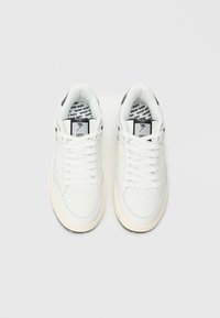 Λευκά αθλητικά παπούτσια με λείο δερμάτινο επάνω μέρος, διάτρητη μύτη, επίπεδα κορδόνια και μαύρες λεπτομέρειες στη φτέρνα. Διαθέτουν ενσωματωμένο εσωτερικό πάτο με το λογότυπο της εταιρείας.