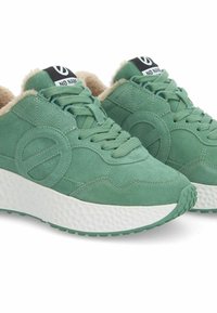Baskets en suède vert avec une doublure beige duveteuse, présentant une semelle blanche texturée, une forme arrondie et un logo circulaire bien en vue sur le côté.