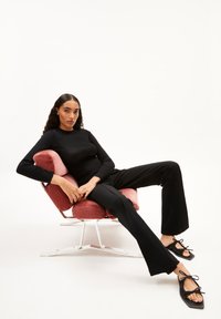 Schwarzes, geripptes Langarmoberteil, schwarze, ausgestellte Hose und schwarze Schnürsandalen, getragen von einem Modell, das auf einem pinken, plüschigen Sessel im Mid-Century-Modern-Stil sitzt.