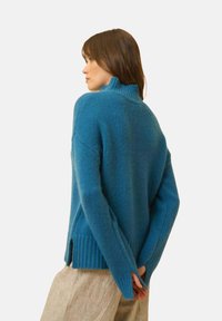 Niebieski sweter dzianinowy z wysokim kołnierzem, ściągaczami w rękawach i bocznymi rozcięciami. Miękka tekstura, luźny krój i jednolity kolor.