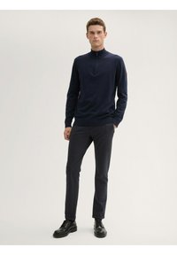 Marineblauer Halbzip-Pullover in schmaler Passform, aus einem glatten Stoff gefertigt. Kombiniert mit dunkel strukturierten Hosen und schwarzen Schnürschuhen.