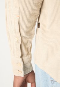 Gros plan sur une manche de chemise en velours côtelé beige avec des boutons et une petite étiquette noire portant la mention BOSS, portée avec un pantalon en jean bleu clair.