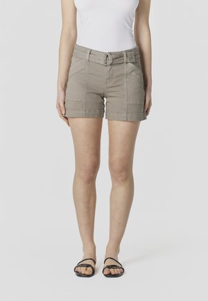 Vrouw met beige shorts met riem, een wit mouwloos topje en zwarte platte sandalen, staande tegen een effen lichte achtergrond.