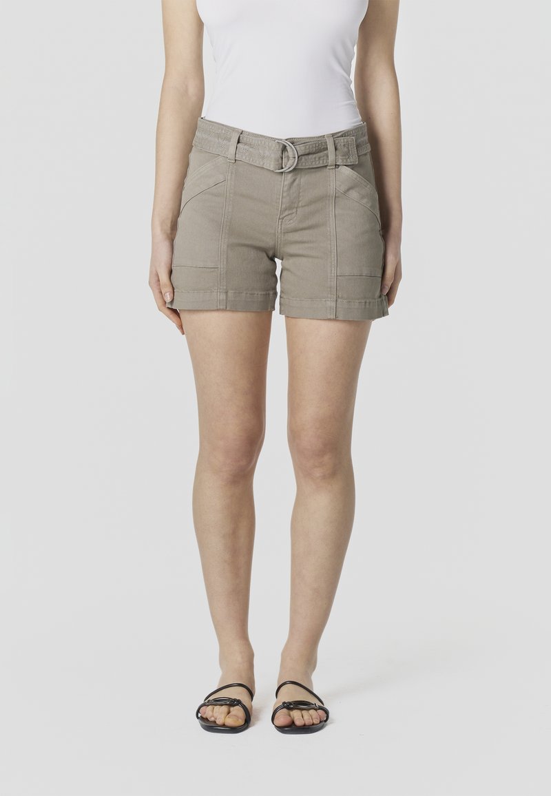 Kvinde iført beige shorts med bælte, hvid ærmeløs top og sorte flade sandaler, stående mod en ensfarvet lys baggrund.
