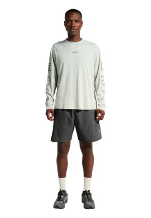Mann steht, trägt ein hellgraues Langarmshirt mit Text, dunkelgraue Shorts, weiße Socken und graue Sportschuhe vor weißem Hintergrund.