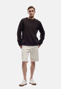 Bruine crewneck sweatshirt, beige shorts en witte sneakers met bruine accenten. Model staat met de handen langs het lichaam in een neutrale houding.