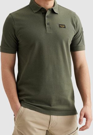 Homme portant un polo vert olive à manches courtes avec un petit logo sur la poitrine et un pantalon beige avec une poche zippée.