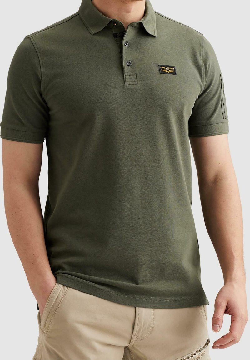 Homme portant un polo vert olive à manches courtes avec un petit logo sur la poitrine et un pantalon beige avec une poche zippée.