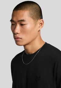 Maglietta nera a maniche lunghe con un piccolo logo ricamato, abbinata a una collana d'argento e un orecchino ad anello. Tessuto morbido e design aderente.