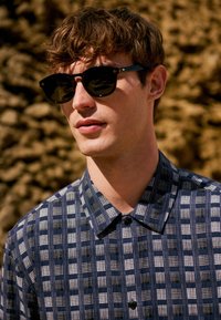 Giovane uomo con occhiali da sole neri e camicia a quadri blu, in piedi all'aperto con sullo sfondo rocce sfocate.