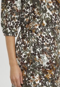 Ziedu raksturā kleita baltā, zaļā un brūnā krāsā; ar elastīgiem piedurkņu galiem un jostu vidukļa regulēšanai. Tekstūrēts audums.