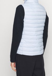 Personne portant un gilet matelassé sans manches bleu clair par-dessus une chemise noire à manches longues et un pantalon noir, vue de dos et de la taille vers le haut.