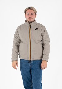 Beżowa lekka kurtka z kołnierzem typu stand-up, pomarańczowym zamkiem błyskawicznym oraz dwoma kieszeniami z przodu. Materiał wydaje się gładki, a krój jest luźny.