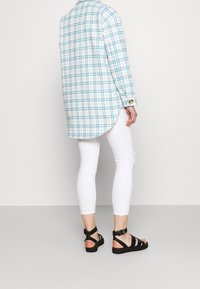 Długa koszula w kratę w białym i turkusowym kolorze, zestawiona z białymi legginsami i czarnymi sandałkami na paski. Gładki materiał; koszula ma zapięte mankiety i zaokrąglony dół.