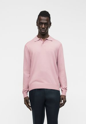 Giovane uomo con indosso una polo a maniche lunghe rosa chiaro e pantaloni scuri, in piedi davanti a uno sfondo grigio chiaro semplice.